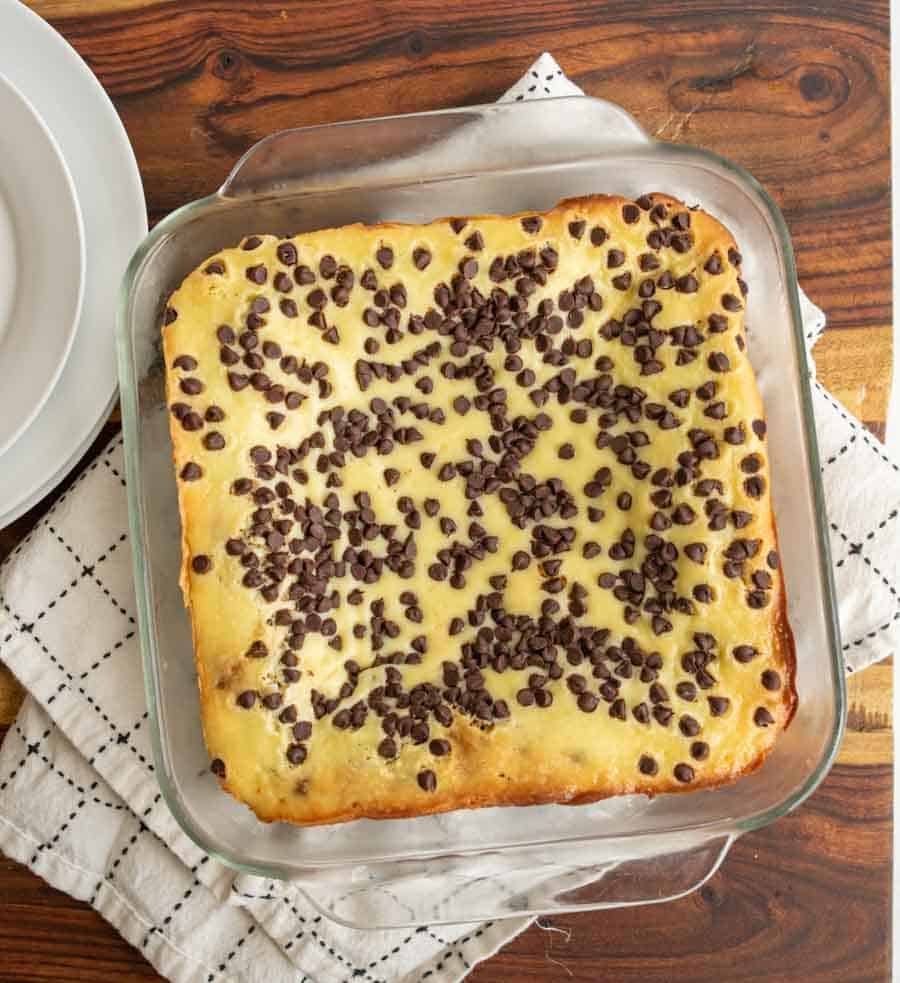 A square glass baking dish holds a baked dessert topped with melted chocolate chips, sitting on a white kitchen towel on a wooden table. Plates are partially visible beside it.