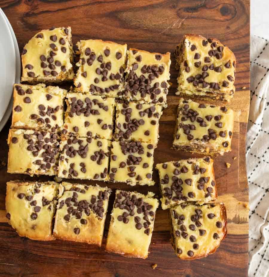 Sliced cookie bars topped with chocolate chips are arranged on a wooden board, with some pieces slightly separated and part of a plate and a white cloth visible at the edges.