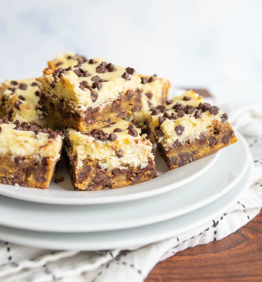 Five dessert bars with chocolate chips and a creamy topping are stacked on a white plate, sitting on a cloth-covered wooden surface.