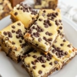 A plate of chocolate chip cheesecake bars stacked on top of each other, featuring a creamy top layer with mini chocolate chips and a dense, brownie-like base.