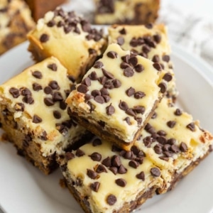 A plate of chocolate chip cheesecake bars stacked on top of each other, featuring a creamy top layer with mini chocolate chips and a dense, brownie-like base.