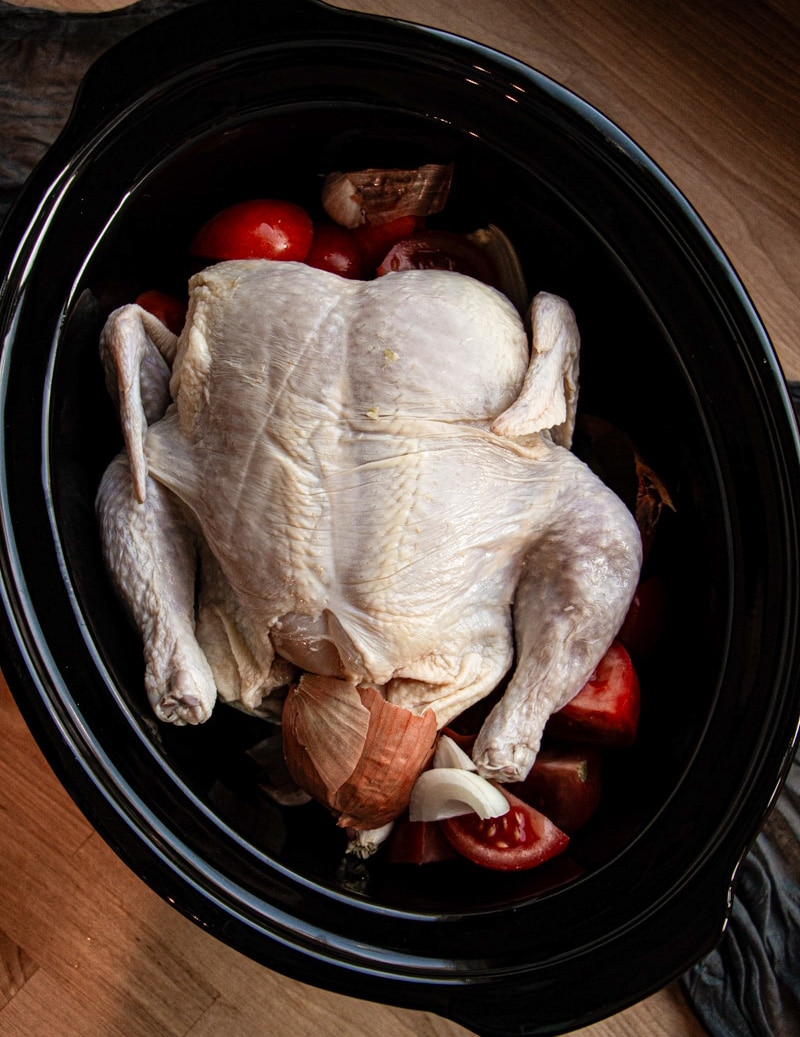 A raw whole chicken is placed in a black slow cooker with sliced tomatoes, onion, and garlic around it, ready for cooking. The image is taken from above on a wooden surface.