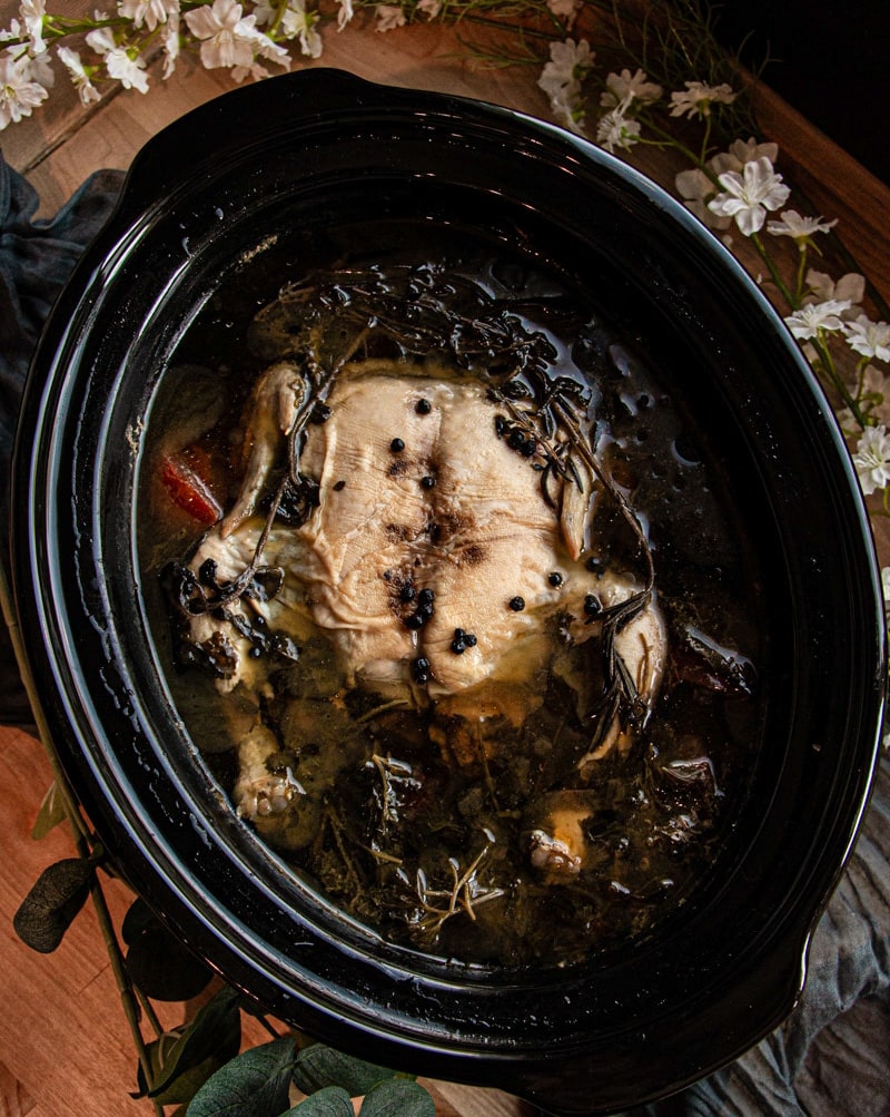 A whole chicken with herbs and spices cooks in broth inside a black slow cooker, surrounded by white flowers and greenery on a wooden table.