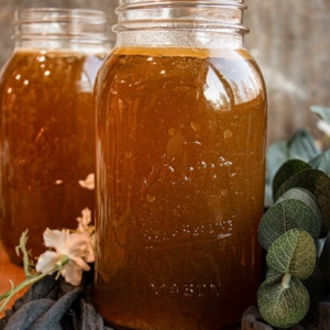 A mason jar filled with brown liquid sits on a cloth, with flowers and greenery nearby. Another filled jar is visible in the background.