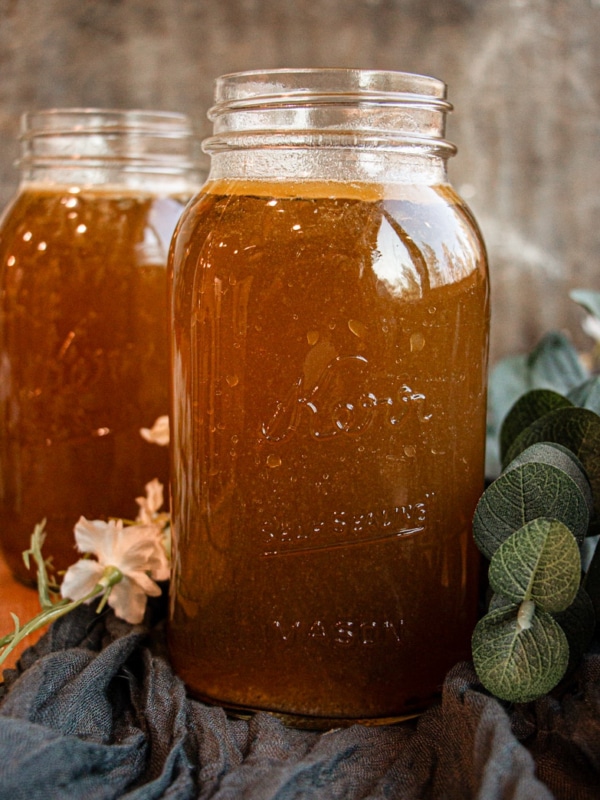 A mason jar filled with brown liquid sits on a cloth, with flowers and greenery nearby. Another filled jar is visible in the background.