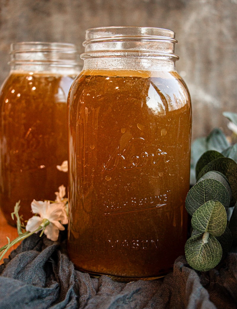 A mason jar filled with brown liquid sits on a cloth, with flowers and greenery nearby. Another filled jar is visible in the background.