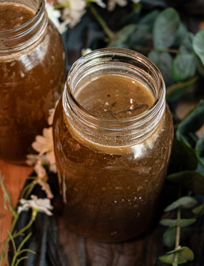 A mason jar filled with a dark brown liquid, possibly herbal tea or broth, sits on a wooden surface surrounded by green leaves and white flowers.