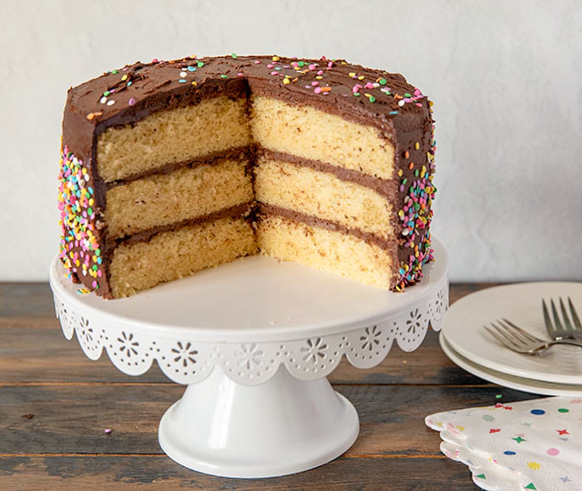 A three-layer yellow cake with chocolate frosting and colorful sprinkles, displayed on a white cake stand. A large slice is missing, revealing the inside layers. Plates and forks are nearby on a wooden table.
