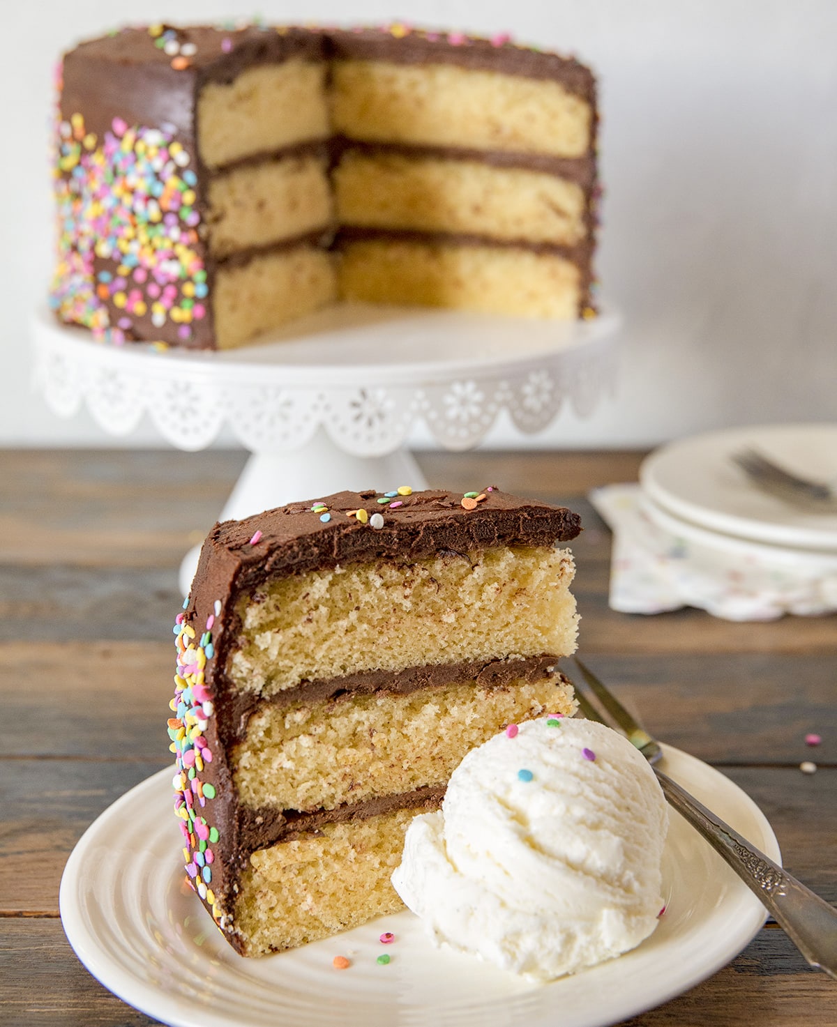 A slice of three-layer vanilla cake with chocolate frosting and sprinkles is served on a plate with a scoop of vanilla ice cream. The rest of the cake is on a stand in the background.