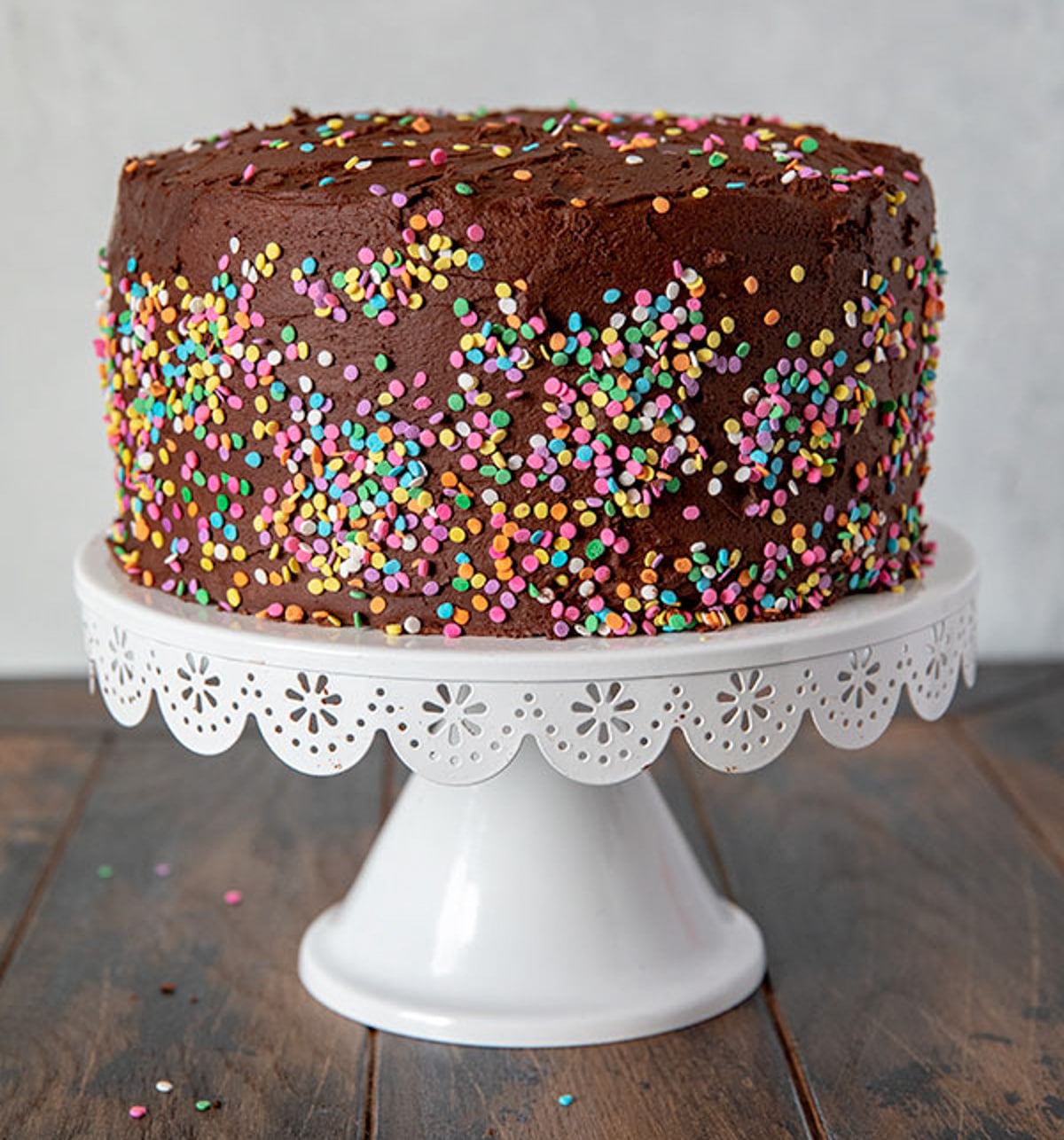 A chocolate frosted layer cake covered with colorful round sprinkles sits on a white decorative cake stand atop a wooden surface.